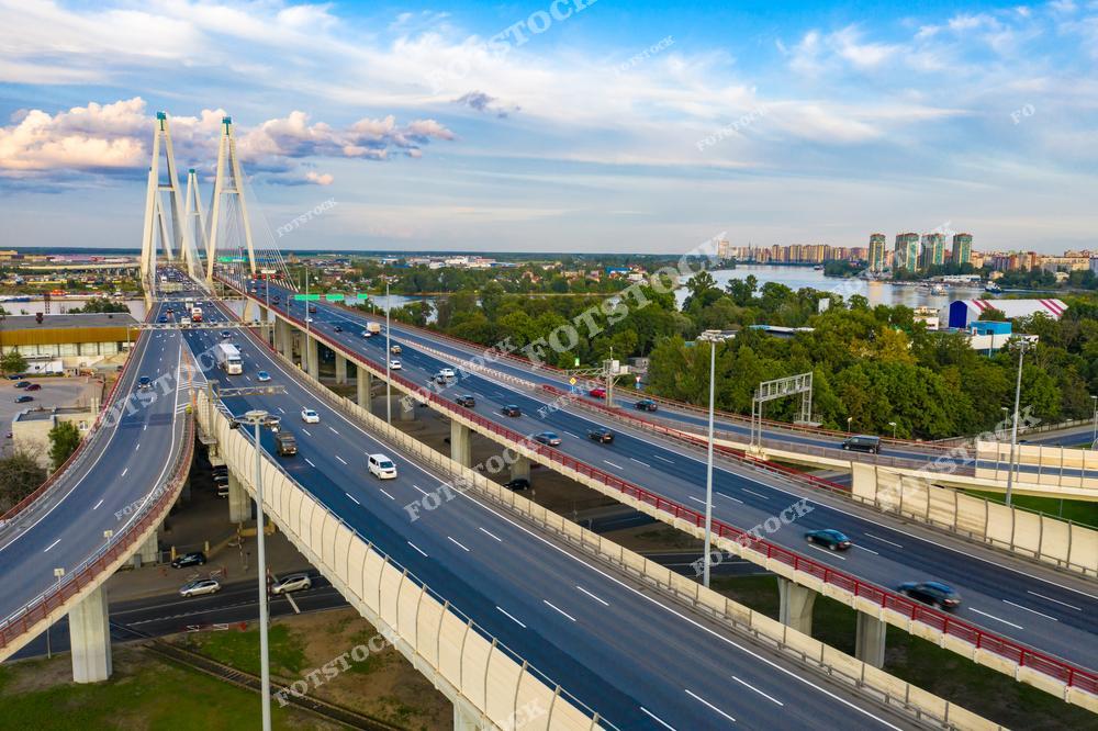 На переднем плане изображен Вантовый мост на Западном скоростном диаметре (ЗСД), являющийся одной из главных транспортных артерий Санкт-Петербурга. Мост выполнен в современном архитектурном стиле с высокими пилонами и вантовыми тросами. Он соединяет южные и северные районы города, проходя через Неву и промышленные зоны. На заднем плане виднеется зелёная зона и новые жилые районы у воды.